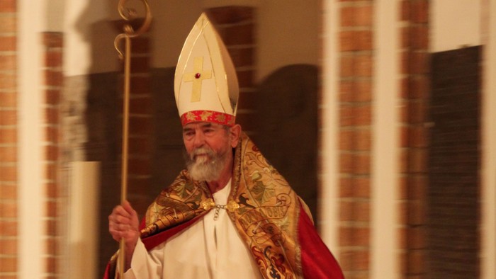 Ein Mann in kirchlicher Kleidung hält einen Stab und steht vor einem Ziegelgebäude.