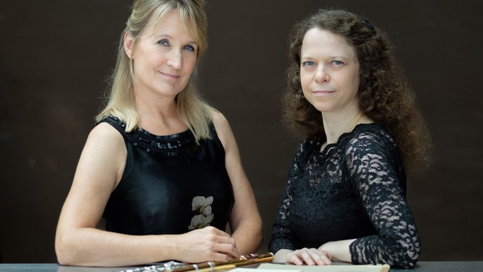 To damer sidder ved bord og ser i kameraet.
