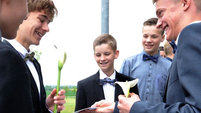 En gruppe unge mænd i formelt tøj samles og smiler, mens de holder blomster og papirer.