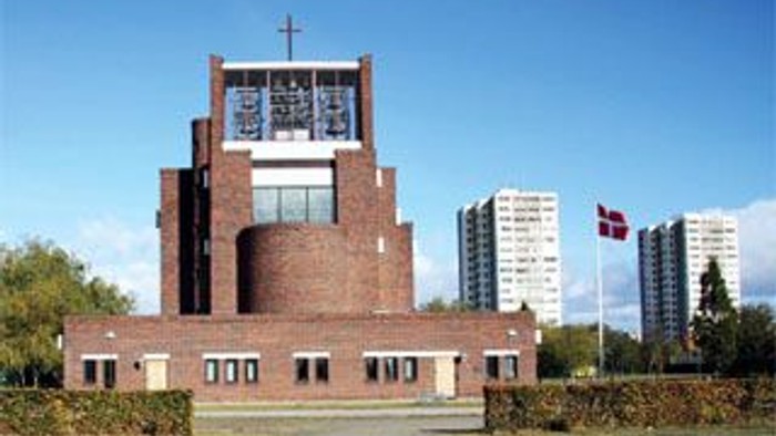 En moderne kirke med tårn og et kors på toppen, placeret i en bymiljø med højhuse og et flagstang.