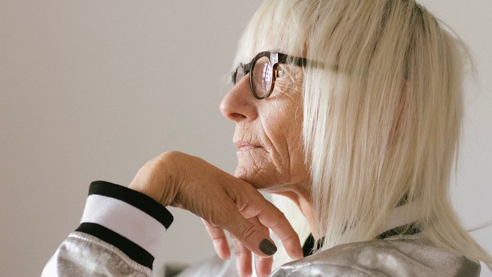 Eine ältere Frau mit Brille und blondem Haar sitzt nachdenklich da.