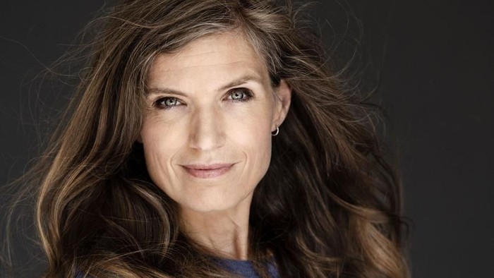 En kvinde med langt, brunt hår, skærer armene og smiler til kameraet.