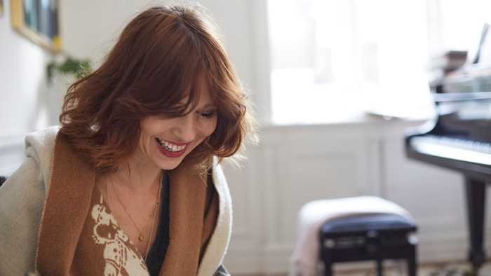 En kvinde med rødt hår sidder ved et piano og smiler.