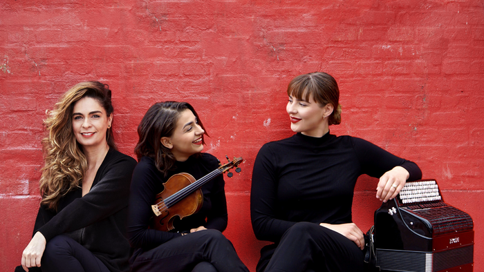 Tre kvinder sidder sammen mod rød mur, to af dem med musikinstrumenter
