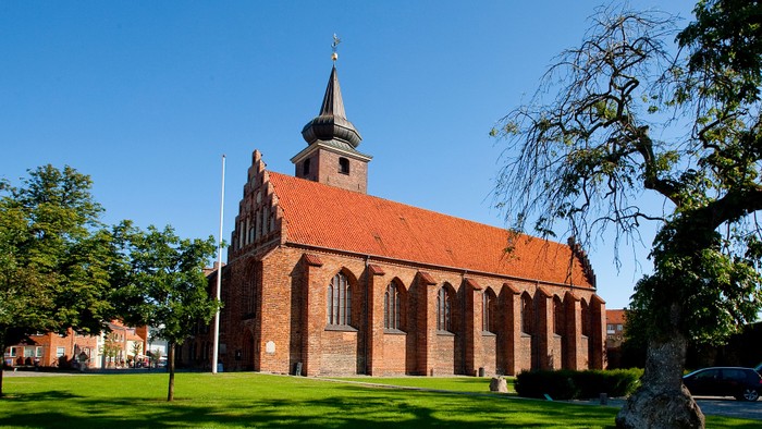 En rød murstenskirke med spir med tårn og grønne træer i forgrunden
