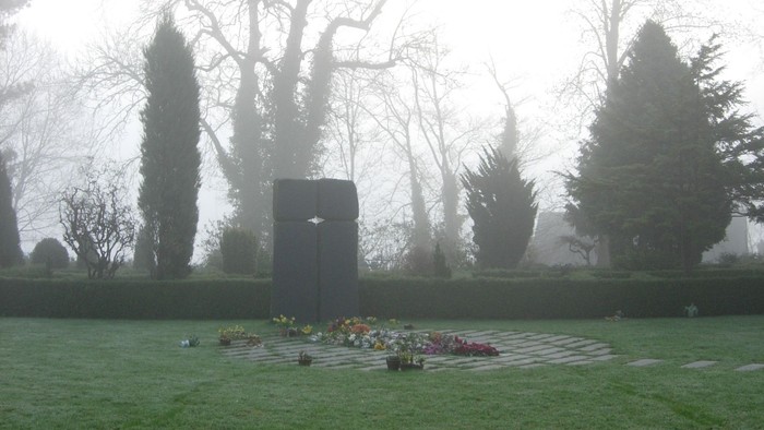 En mindeplads med blomster og en høj monolitisk sten i en trægt og tåge.