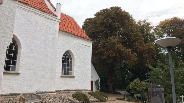 Del af en hvid kirke med rødt tag og efterårsgrønne træer i baggrunden