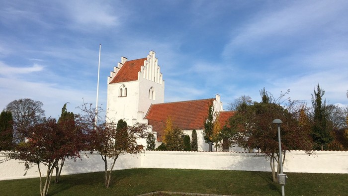 Et hvidt kirkebygning med rødt tag og tårn, omgivet af træer og en græsplæne