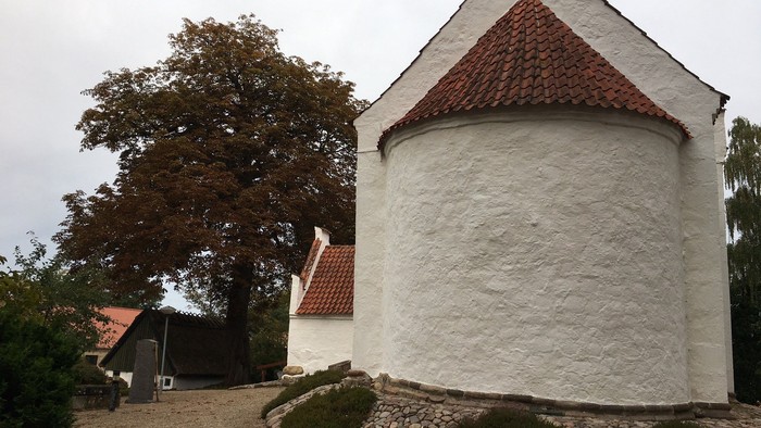 Del af hvidkalket kirke med rødt tag og et stort træ i baggrunden