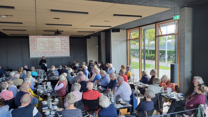 En stor gruppe af ældre mennesker sidder ved bord i et lokalet og spiser.