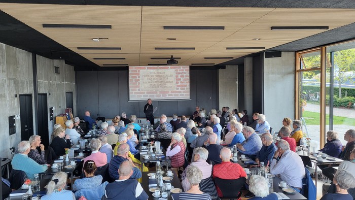 En gruppe af ældre mennesker sidder ved bord i et lokalet og lytter til en foredragsholder.