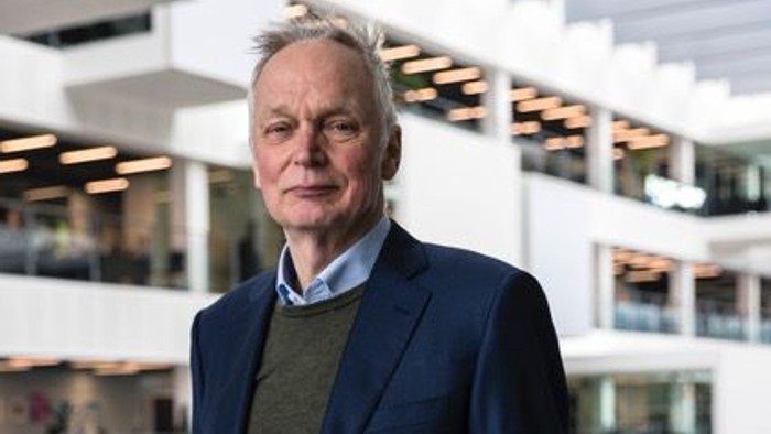 En eldre mand i blå jakkesæt og grøn pullover står i et moderne indkøbscenter.