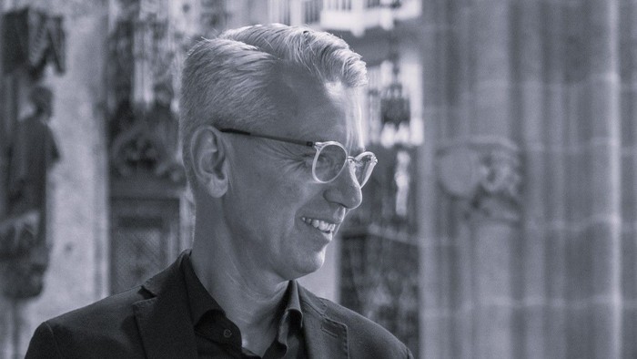 Ein älterer Mann mit Brille lächelt in einem Raum mit steinernen Verzierungen.