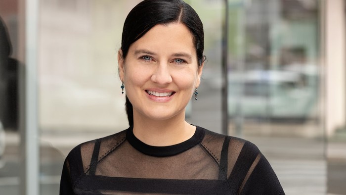 Eine Frau mit dunklem Haar lächelt in die Kamera, sie trägt ein schwarzes Oberteil.