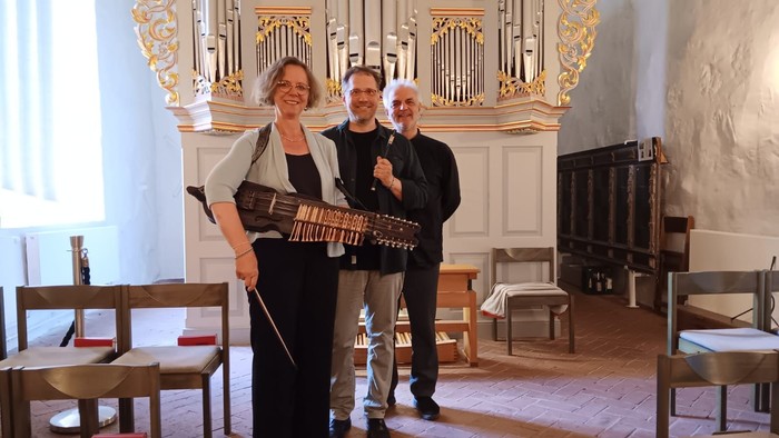 Drei Personen posieren vor einem Kirchenorgel, zwei halten Instrumente
