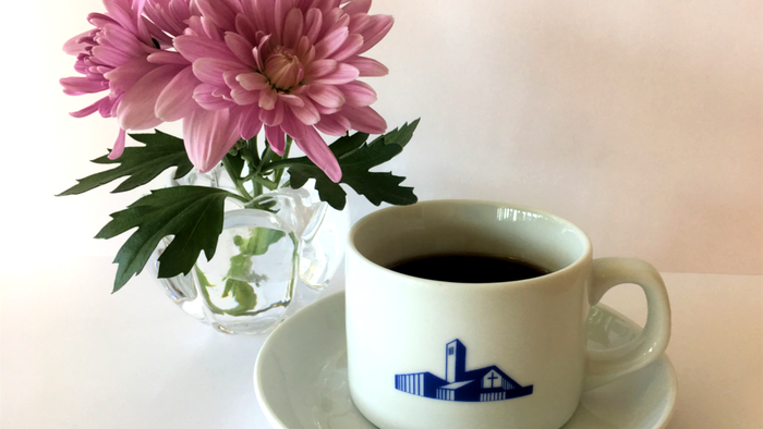 En kop kaffe med et bygningslogo står ved siden af en vase med livlige lyserøde blomster på en underkop.