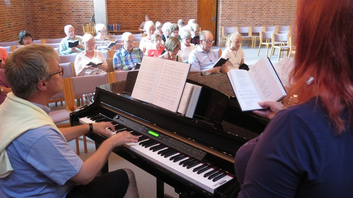 En gruppe af mennesker deltager i en sangprove, hvor en mand spiller piano og en kvinde holder partituret op.
