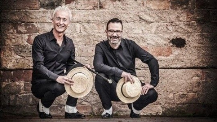 Zwei Männer posieren mit Hüten vor Ziegelmauer.