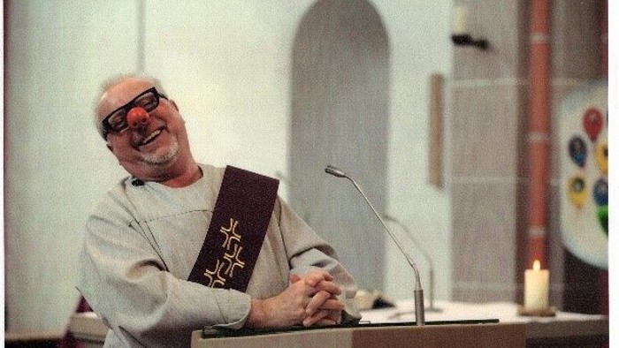 Mann steht lachend hinter Pult mit Mikrofon und Buch