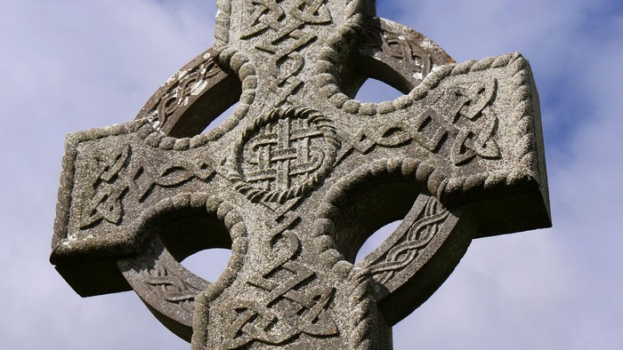 En udadret stenkors med intricate knoting og mønstre, stående mod et klart blå himmel.