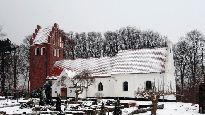 En snedækket kirke med et rødt tårn og et hvidt tag, omgivet af bærbare træer og sne.