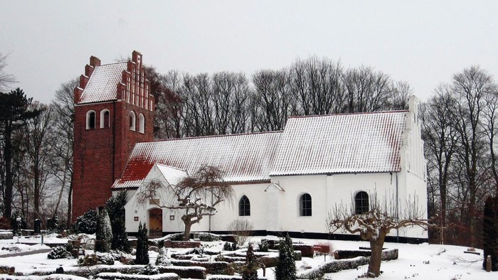 En snedækket kirke med et rødt tårn og et hvidt tag, omgivet af bærbare træer og sne.