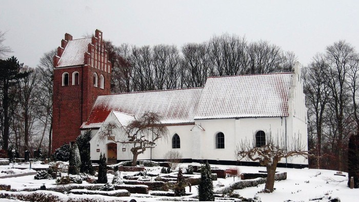 En snedækket kirke med et rødt tårn og et hvidt tag, omgivet af bærbare træer og sne.