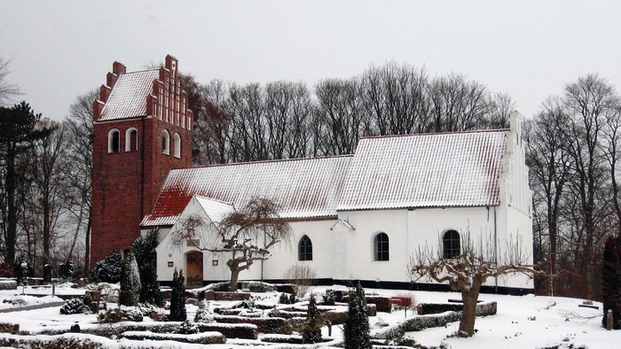 En snedækket kirke med et rødt tårn og et hvidt tag, omgivet af bærbare træer og sne.