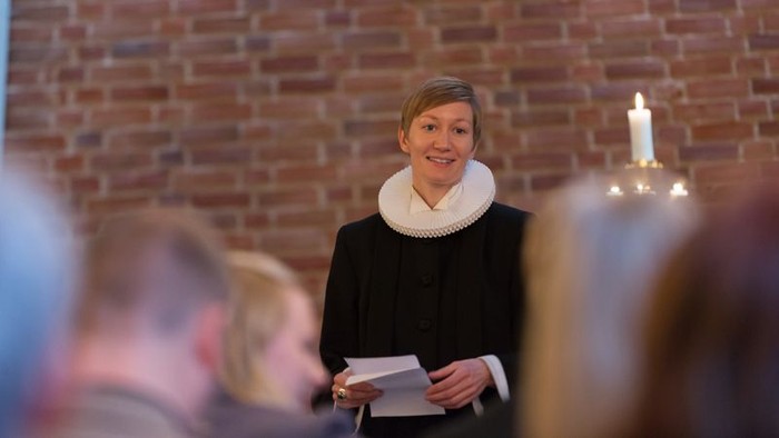 En kvinde i sort jakke og hvid skærm stander foran en mur med lysende lys og holder en bog.