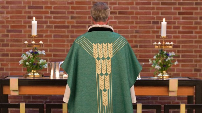 Mand i grøn liturgisk kappe står for alter med lysstaker og blomster.