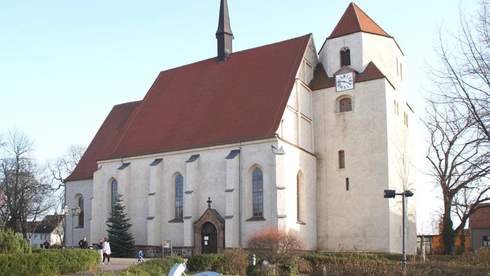Weiße Kirche mit rotem Dach und hohem Turm, umgeben von Bäumen und Büschen
