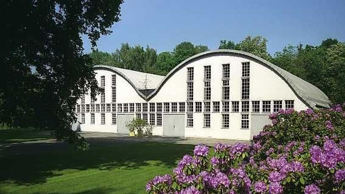 Ein großes weißes Gebäude mit gewölbtem Dach und vielen Fenstern, umgeben von grünem Gras und blühenden lila Sträuchern unter einem klaren blauen Himmel.