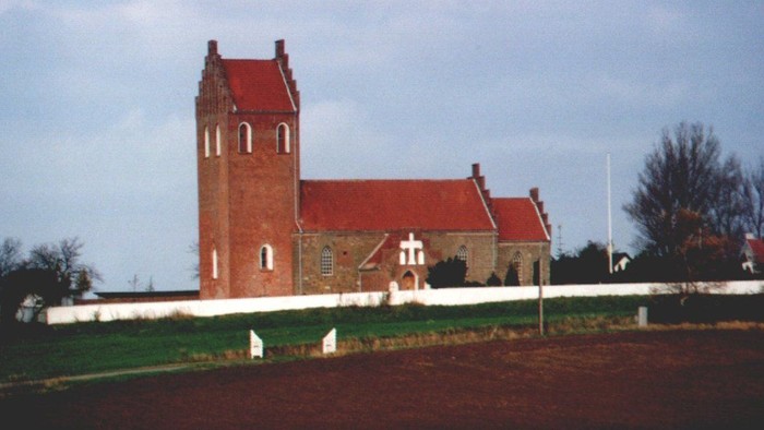 En rød kirke med tårn og hvide omgivelser, beliggende på en grøn og rød mark med træer i baggrunden