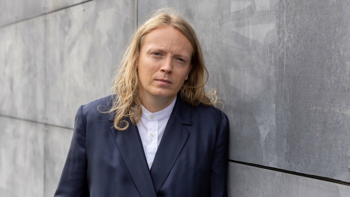 Mand med lang blond hår og mørkt jakkesæt står ved betonvej.