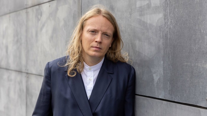 Mand med lang blond hår og mørkt jakkesæt står ved betonvej.