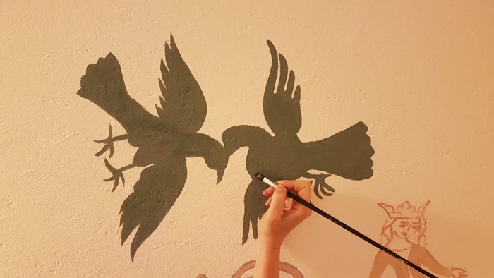 En hånd holder en stang med to papirflueklip, mens en murmalet figur ser til.