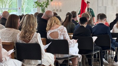 En formel forsamling med deltagere siddende og en taler, der holder et rødt flag forrest.