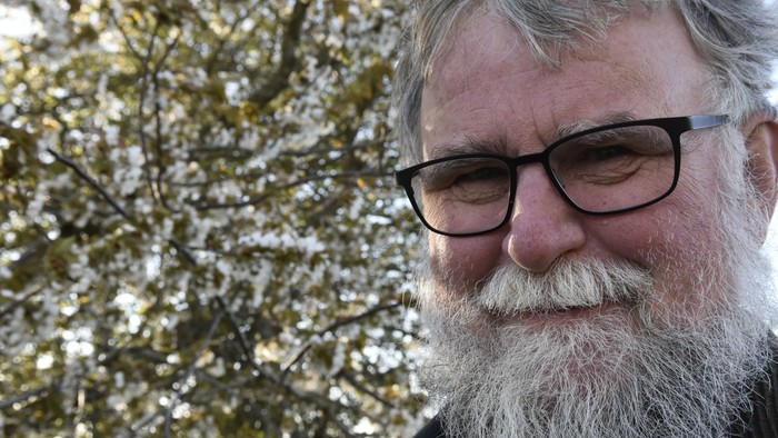 En bjergtegnende mand med briller og skæg står foran et træ med blomster.