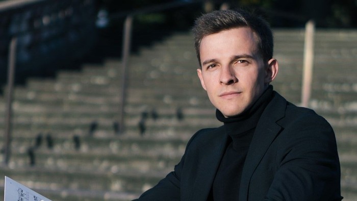 Junger Mann in Anzug sitzt in antiker Arena und hält Noten.