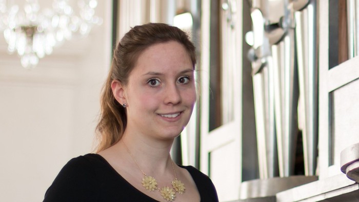 Eine junge Frau mit blondem Haar und schwarzem Oberteil lächelt in einem Raum mit Kronleuchter.