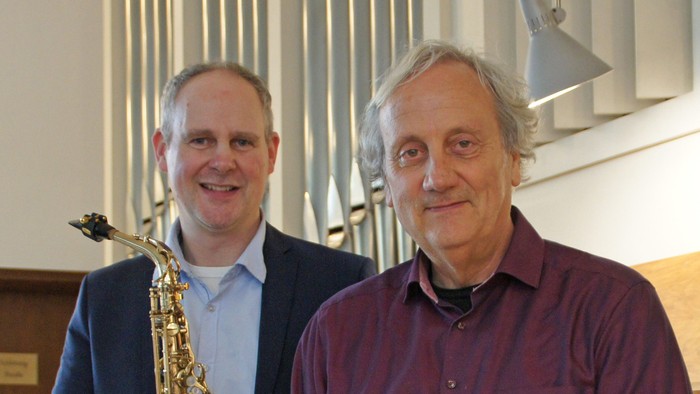 Zwei Männer posieren vor einem Orgelpfeifen, einer hält eine Saxophon