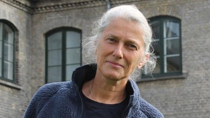 En eldre mand med hvidt hår står foran et murstenshus med mange vinduer.