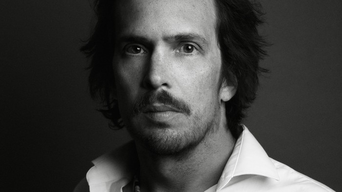 Mand med langt hår og skæg, der bærer en halskæde og et hvide skjorte.