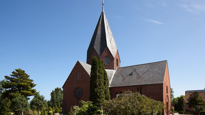 En rød kirke med spids tårn og kryds på toppen, omgivet af træer og buske under et klarblåt himmel.