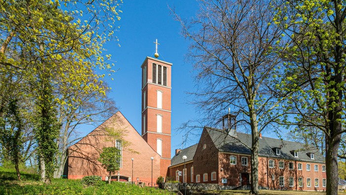 Kirchturm aus rotem Backstein mit Kreuz, umgeben von Bäumen und grünem Gras