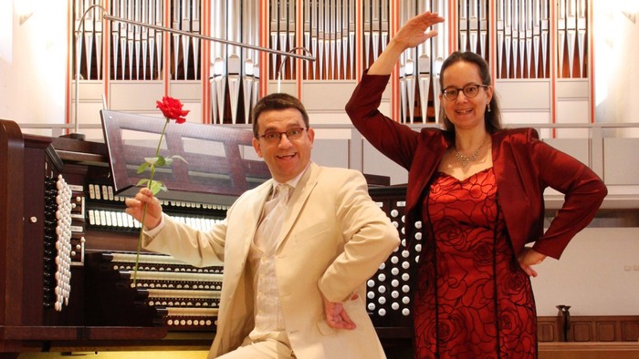 Ein Mann und eine Frau posieren vor einem Kirchenorgel, er hält eine rote Rose.