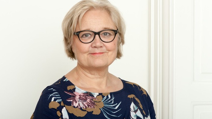 En kvinde med kort, blond hår og briller poserer mod en hvid væg.