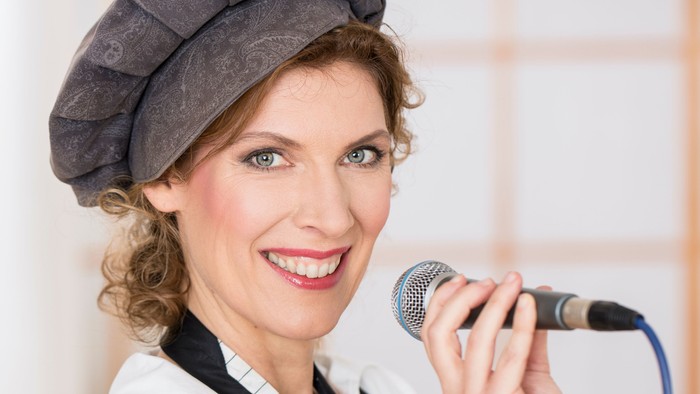 Eine Frau singt in ein Mikrofon und trägt eine weiße Bluse mit schwarzer Schleife und eine graue Mütze.