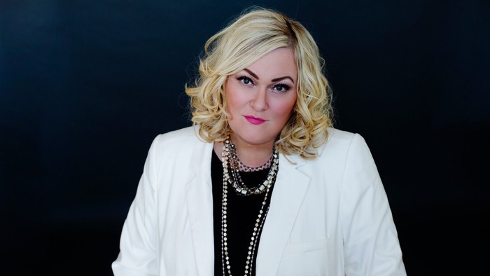 En kvinde med blond hår og røde lipper bærer et hvidt jakkesæt og mørke smykker.