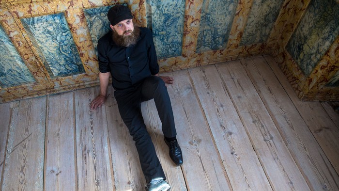 Mand med lang barbe sidder på gulv i lille rum med blå og gule mønstre på vægge